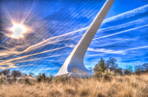 A striking white bridge tower against a blue sky with wispy clouds and sun rays, surrounded by tall grass and trees.