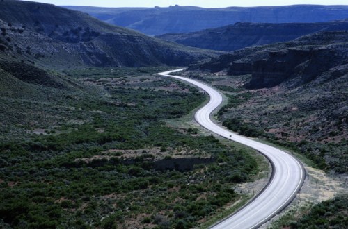 A winding road cuts through a lush green valley surrounded by rocky hills and distant mountains under a clear blue sky.