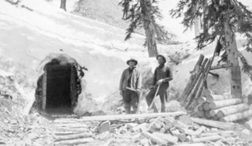Two men stand near a snow-covered mine entrance, surrounded by logs and trees in a historical black-and-white photo.