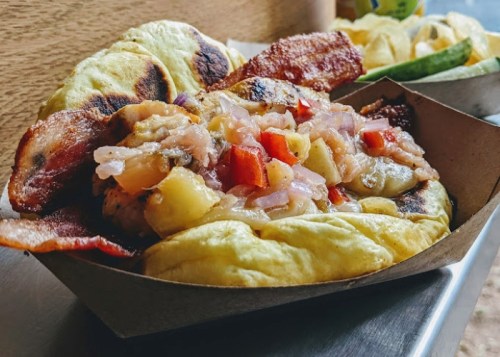 A close-up of a food dish featuring grilled bacon, sautéed vegetables, and a soft bread wrap in a paper tray.