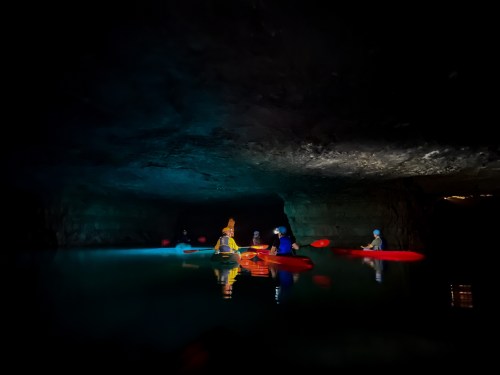 A group of kayakers paddles in a dark cave, illuminated by colorful lights reflecting on the water's surface.