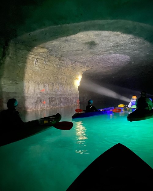 Kayakers explore a dimly lit cave with glowing water, illuminated by a beam of light and colorful kayak paddles.