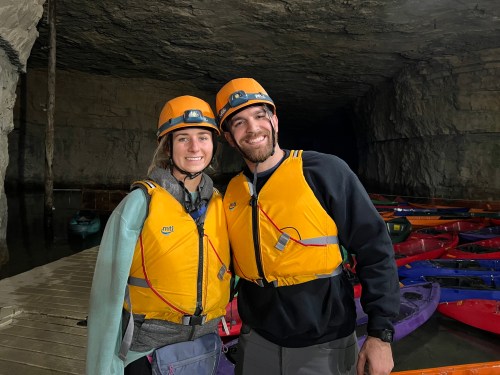 Two people in bright orange helmets and life jackets smile in a cave with kayaks in the background.