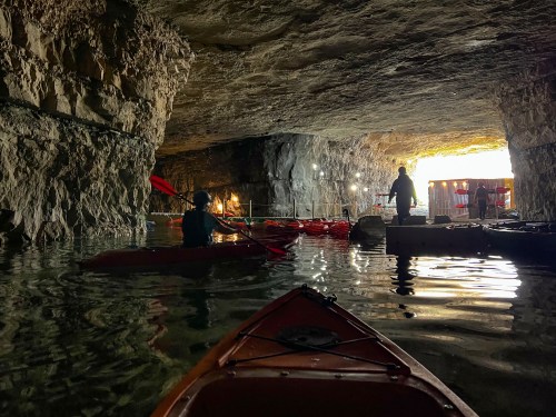 Two kayakers explore a cavern with rocky walls, illuminated by soft light, reflecting on the water's surface.