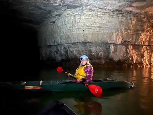 A person in a kayak wearing a helmet and life jacket paddles in a dark cave with illuminated rock walls.