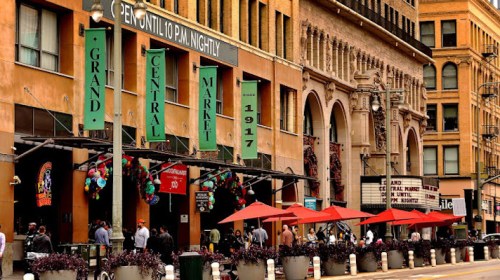 A bustling market entrance with colorful signage, outdoor seating, and people gathered under red umbrellas.