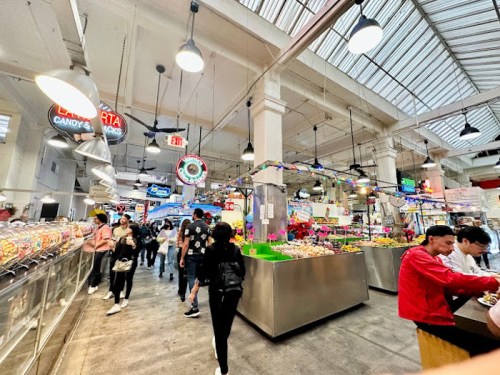 A bustling market interior with colorful stalls, people shopping, and bright signage overhead.