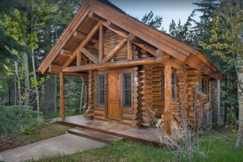 A rustic log cabin surrounded by trees, featuring a wooden porch and large front windows.