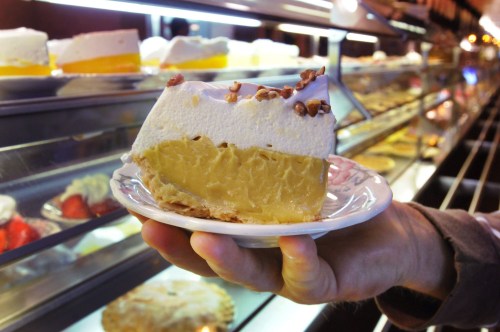 A slice of creamy dessert with a graham cracker crust, topped with whipped cream and nuts, held in a hand.