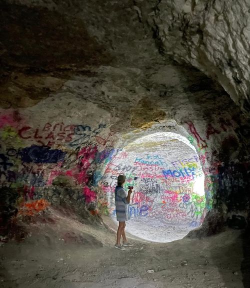 A person stands inside a cave, illuminated by light, surrounded by colorful graffiti on the walls.