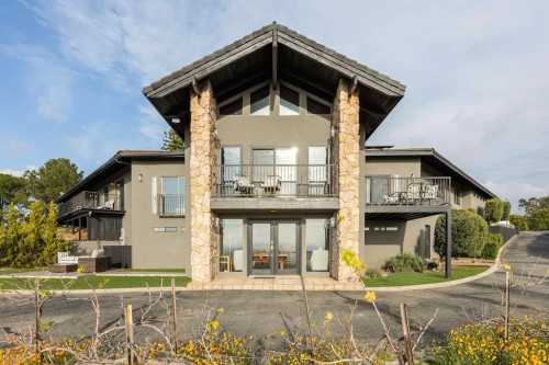 Modern two-story house with stone accents, large windows, and balconies, surrounded by greenery and a winding driveway.