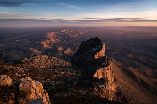 A dramatic mountain peak rises above a vast, rolling landscape at sunset, casting long shadows across the terrain.