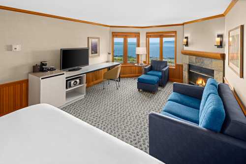 Cozy hotel room with a blue sofa, armchair, desk, TV, and a view of the ocean through large windows.