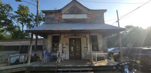 A rustic, weathered store with a Coca-Cola sign, rocking chairs on the porch, and a sunny backdrop.