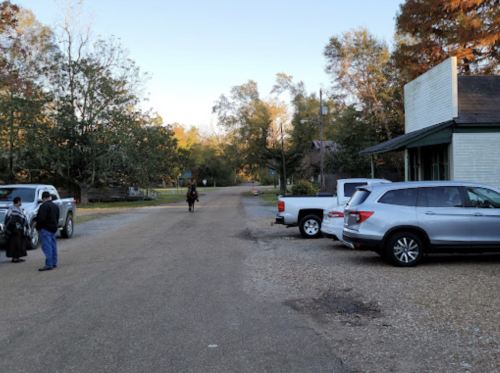 A quiet rural road with parked vehicles and people near a building, surrounded by trees and autumn foliage.