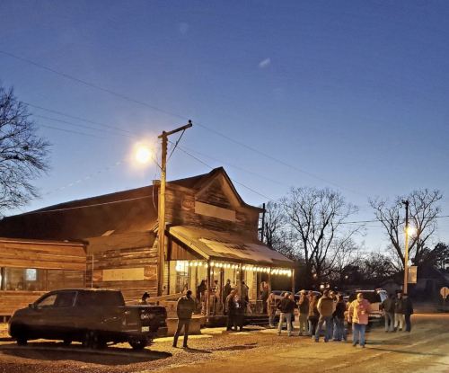 A crowd gathers outside a rustic building at dusk, with warm lights glowing and a clear blue sky above.