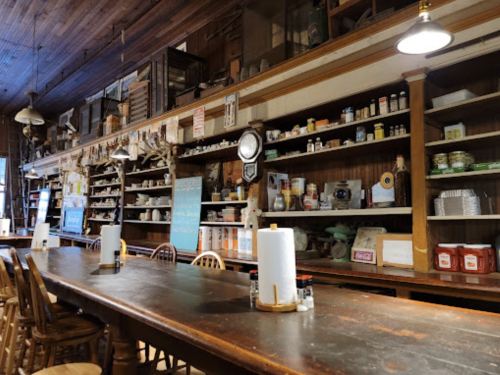 A rustic dining area with a long wooden table and shelves filled with jars, spices, and kitchenware.