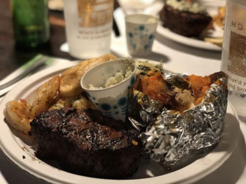A plate of food featuring steak, shrimp, baked potato, and a small cup of mashed potatoes, with drinks in the background.