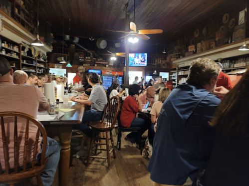 A bustling restaurant interior with patrons seated at tables and a bar, featuring wooden decor and a TV in the background.