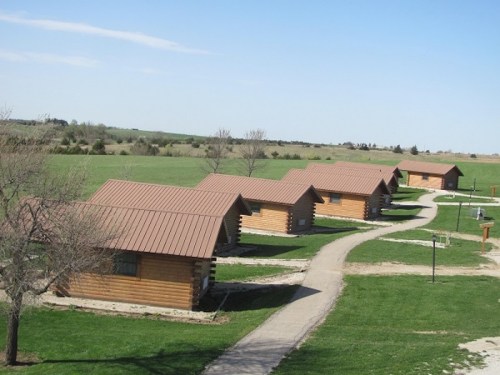 A row of log cabins with metal roofs, set on a grassy landscape under a clear blue sky.