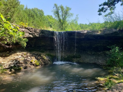 A serene waterfall cascading over rocky cliffs into a calm pool, surrounded by lush greenery under a clear blue sky.