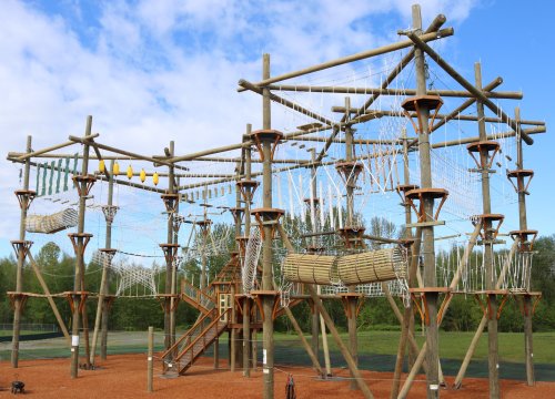 A large wooden climbing structure with ropes and nets, set in a grassy area under a blue sky.