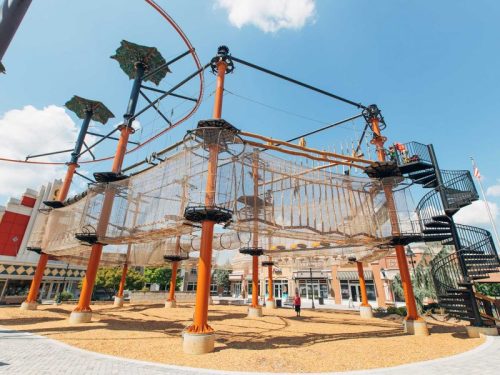 A large outdoor climbing structure with nets and a spiral staircase, surrounded by a gravel area and buildings in the background.