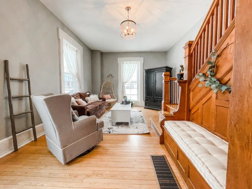 Cozy living room with a gray armchair, wooden staircase, and natural light from large windows.