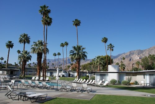 A sunny resort with palm trees, lounge chairs, and modern buildings against a backdrop of mountains and clear blue sky.