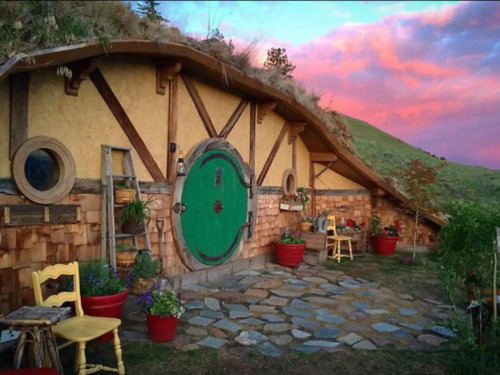 A charming hobbit-style house with a green door, surrounded by colorful flowers and a scenic mountain backdrop at sunset.