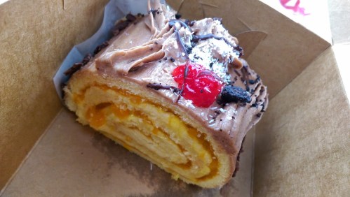 A slice of chocolate frosted roll cake with orange filling, topped with a cherry and chocolate shavings in a takeout box.