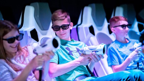 Three kids wearing 3D glasses sit in futuristic seats, holding gaming guns and enjoying an immersive experience.
