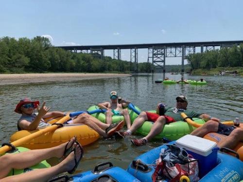 Group of people relaxing on colorful inner tubes in a river, with a bridge and trees in the background on a sunny day.