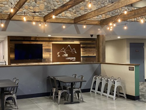 A cozy café interior with wooden beams, string lights, and a sign reading "Cliff Rangers Café." Tables and chairs are arranged.