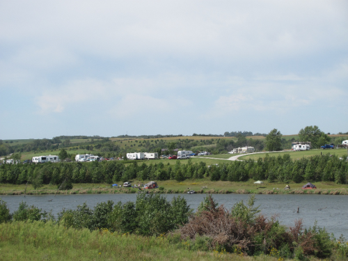 A scenic view of a river with RVs parked along the shore and green fields in the background under a cloudy sky.