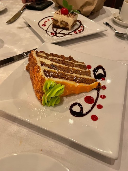 A slice of carrot cake with cream cheese frosting, garnished with green icing and red sauce, on a white plate.