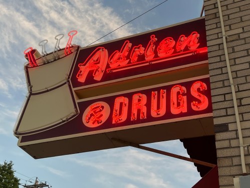 Neon sign for "Adrien Drugs" featuring a beaker design, set against a blue sky.