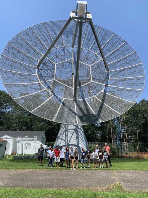 A group of people stands in front of a large satellite dish on a sunny day, surrounded by greenery.