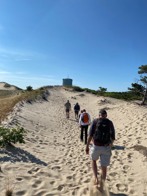 A group of hikers walking on sandy dunes with a water tower in the background under a clear blue sky.