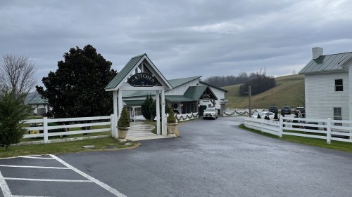 A rustic building with a green roof and white trim, surrounded by a grassy landscape under a cloudy sky.