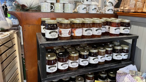 A wooden shelf displaying various jars of preserves and mugs in a cozy shop setting.