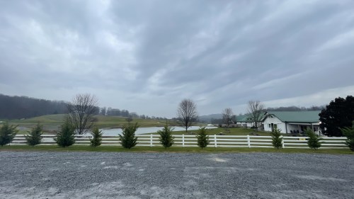A cloudy sky over a green landscape with a pond, white fence, and a house in the distance.