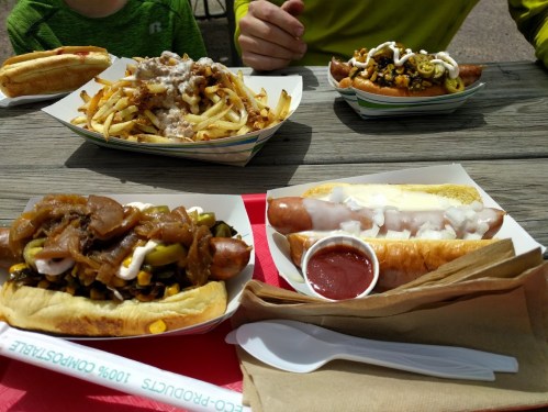 A table with three trays of food: hot dogs with toppings, loaded fries, and condiments on a picnic table.