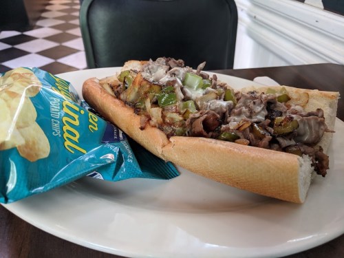 A loaded sandwich with beef, peppers, and onions on a plate, accompanied by a bag of potato chips.