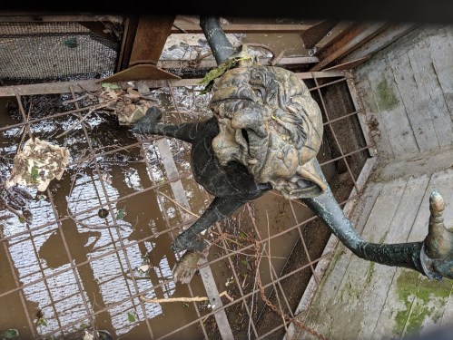 A weathered, ornate metal figure with a lion-like face, partially submerged in muddy water and surrounded by debris.