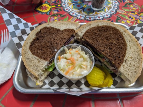 A sandwich on dark bread, cut in half, served with a side of coleslaw and pickles on a colorful table.