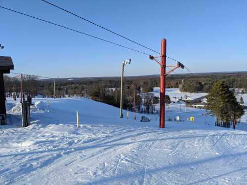 A snowy ski slope with a ski lift, surrounded by trees and a clear blue sky in the background.