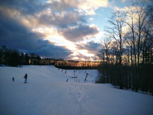 A snowy ski slope under a dramatic sky with clouds and a sunset, surrounded by trees.