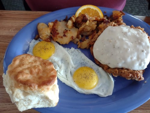A blue plate with fried chicken topped with gravy, two sunny-side-up eggs, hash browns, and a biscuit with an orange slice.