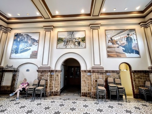 Interior of a historic building featuring large murals and stacked chairs, with a person sitting against the wall.